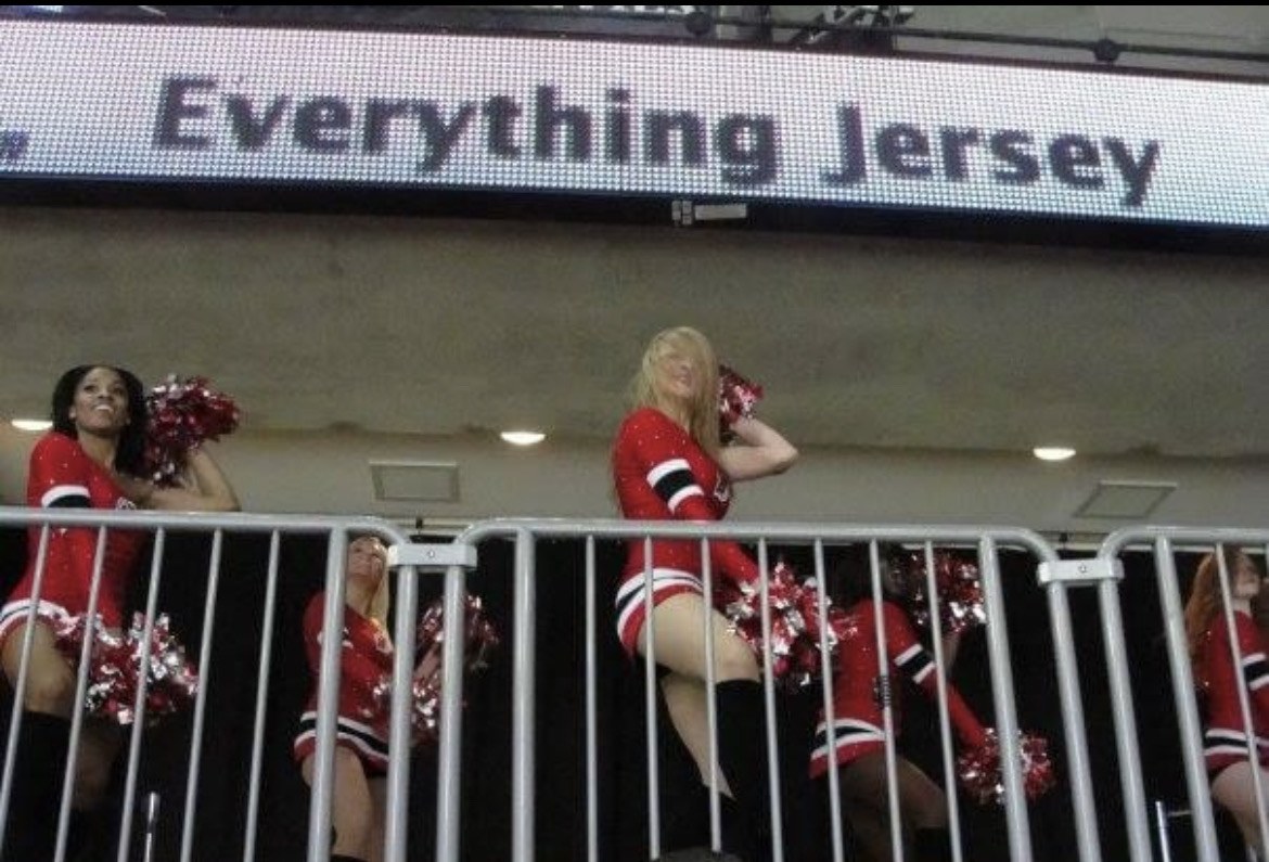 On the New Jersey Devils dance team, pom-poms up, under an Everything Jersey sign.